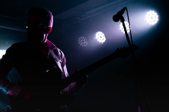 Mann mit Gitarre im Schatten, beleuchtet von bunten Bühnenlichtern.