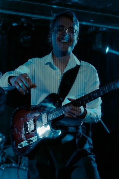 Mann mit Gitarre auf der Bühne, lächelt in schummrigen Lichtern.