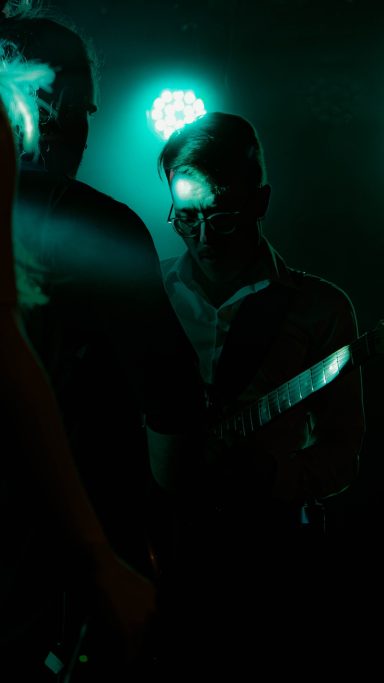 Dunkle Bühne mit Personen, ein Musiker konzentriert mit Gitarre im Vordergrund.