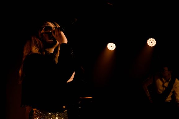 Musikerin auf der Bühne, singend im schwachen Licht mit Scheinwerfern im Hintergrund.
