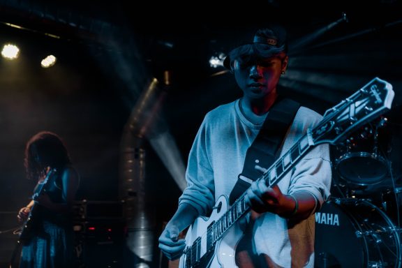 Musiker mit E-Gitarre auf der Bühne, beleuchtet von Bühnenlicht, in aufgeheiterter Atmosphäre.
