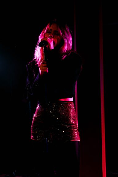Frau mit Mikrofon, trägt einen glitzernden Rock und hat pinkes Licht im Hintergrund.