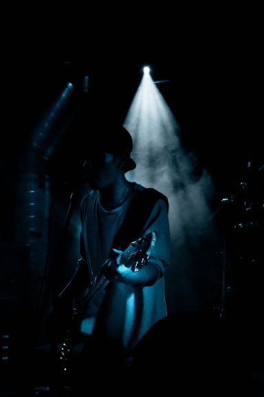 Dunkle Bühnenaufnahme eines Musikers mit Gitarre im Lichtschein.