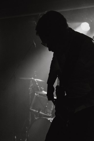 Silhouette eines Musikers, der Gitarre spielt, mit diffusem Licht und Schlagzeug im Hintergrund.