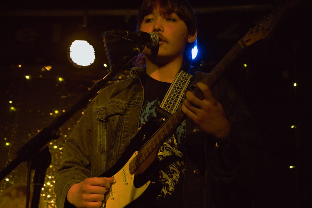 Junger Mann spielt E-Gitarre auf der Bühne, umgeben von bunten Lichtern.