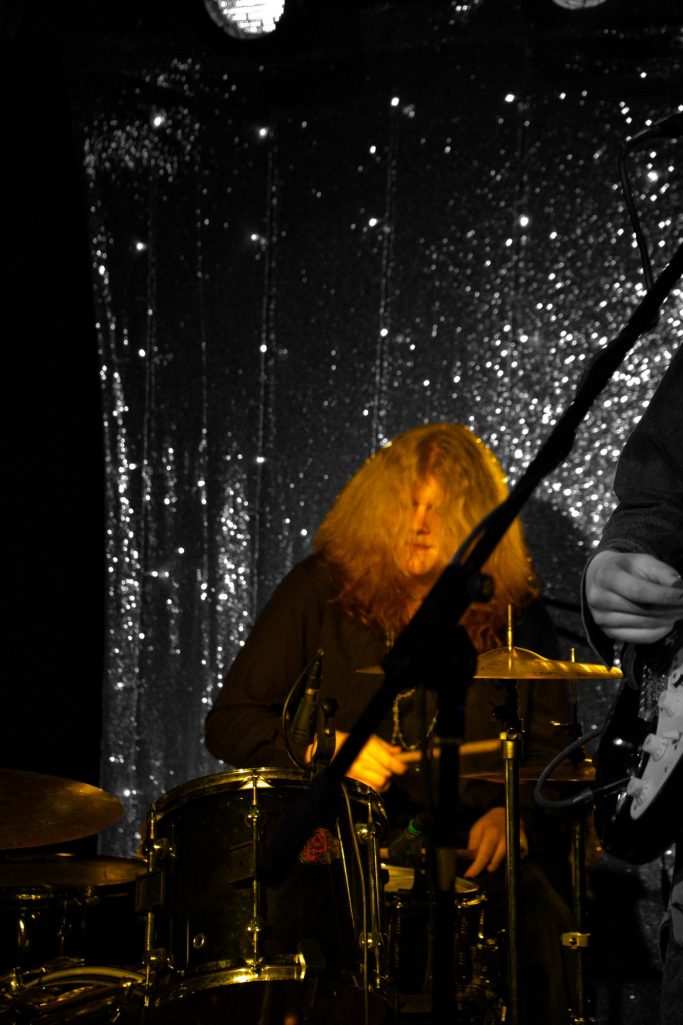 Ein Musiker mit lockigem Haar spielt Schlagzeug vor glitzerndem Hintergrund.