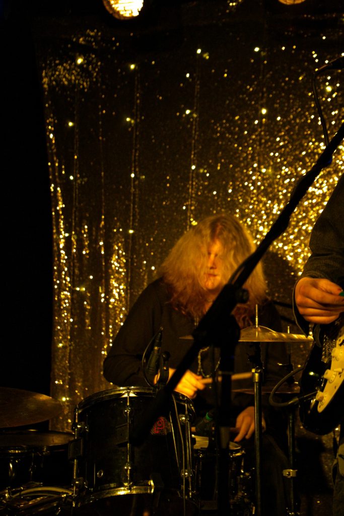 Ein Musiker mit langen, lockigen Haaren spielt Schlagzeug vor einem glitzernden Hintergrund.