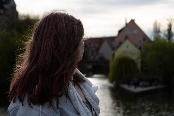 Frau mit langen, roten Haaren blickt auf einen ruhigen Kanal und historische Gebäude.