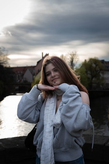 Junge Frau mit langen Haaren posiert lächelnd, im Hintergrund ist eine Landschaft mit Wasser.