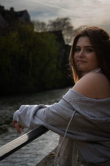Eine junge Frau lehnt an einem Geländer, im Hintergrund fließt ein ruhiger Fluss.