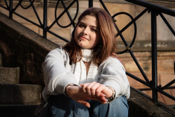 Junge Frau mit langen, roten Haaren, sitzt auf einer Treppe und lächelt.