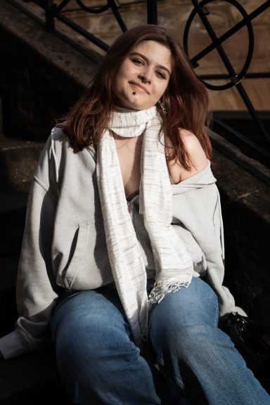 Junge Frau mit langen braunen Haaren trägt einen grauen Hoodie und einen weißen Schal.