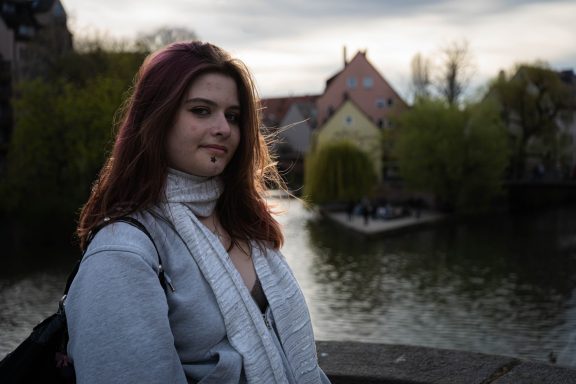 Junge Frau mit braunen Haaren steht an einem Wasserkanal, umgeben von Bäumen und Häusern.