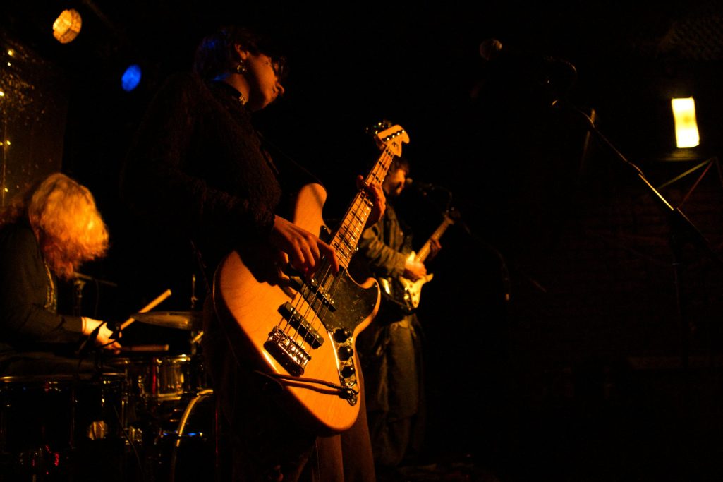 Drei Musiker spielen in einem dunklen Raum, einer mit einer E-Gitarre und zwei mit anderen Instrumenten.