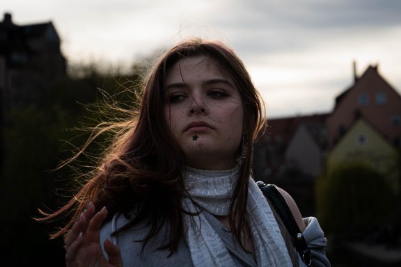 Junge Frau mit langen Haaren und nachdenklichem Ausdruck vor einem wolkenverhangenen Himmel.