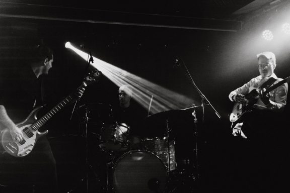Drei Musiker auf der Bühne, mit Gitarre, Bass und Schlagzeug, beleuchtet von Scheinwerfern.