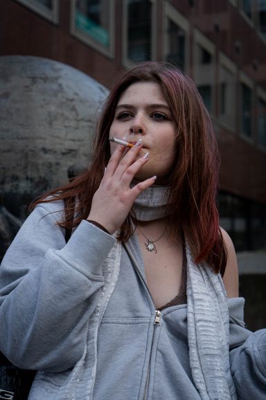Junge Frau mit rotem Haar, die eine Zigarette raucht, in urbaner Umgebung.