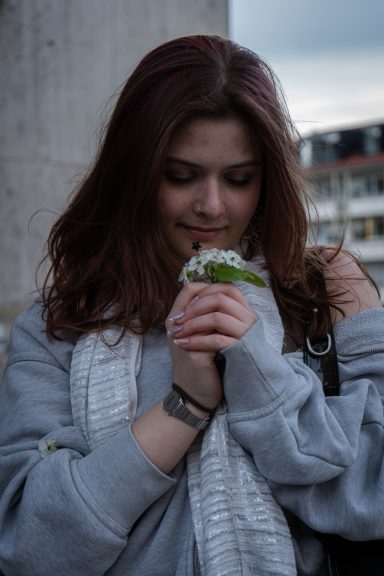 Frau mit langen, braunen Haaren umarmt eine Pflanze, trägt einen grauen Pullover und einen Schal.