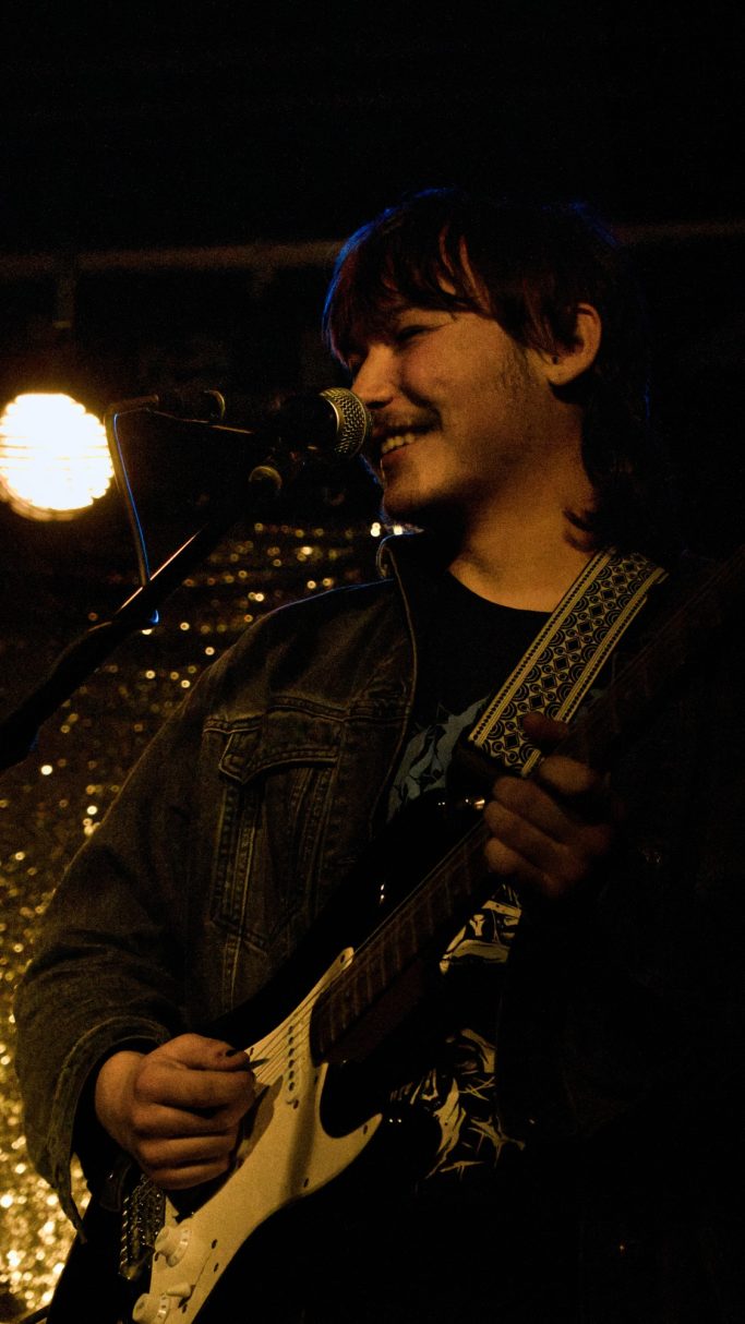 Junger Musiker mit Gitarre, lächelnd auf einer Bühne mit schwachem Licht.