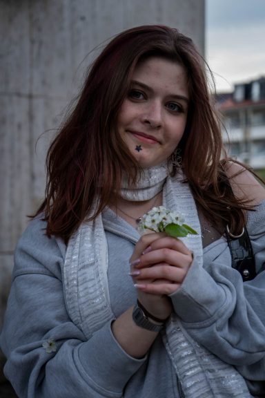 Junge Frau mit langen braunen Haaren, trägt eine graue Jacke und hält eine kleine Pflanze.