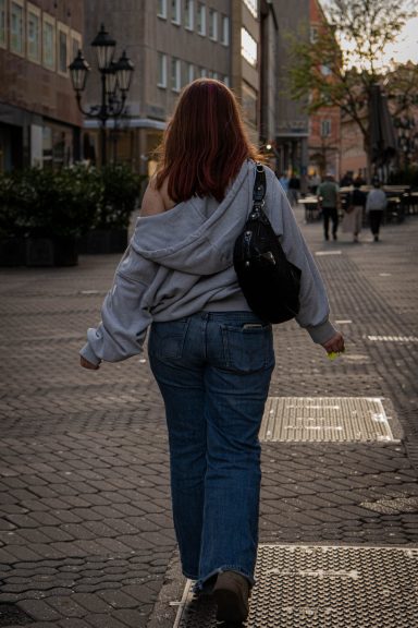 Person mit rotem Haar in Grauem Sweatshirt und Jeans geht eine Straße entlang.