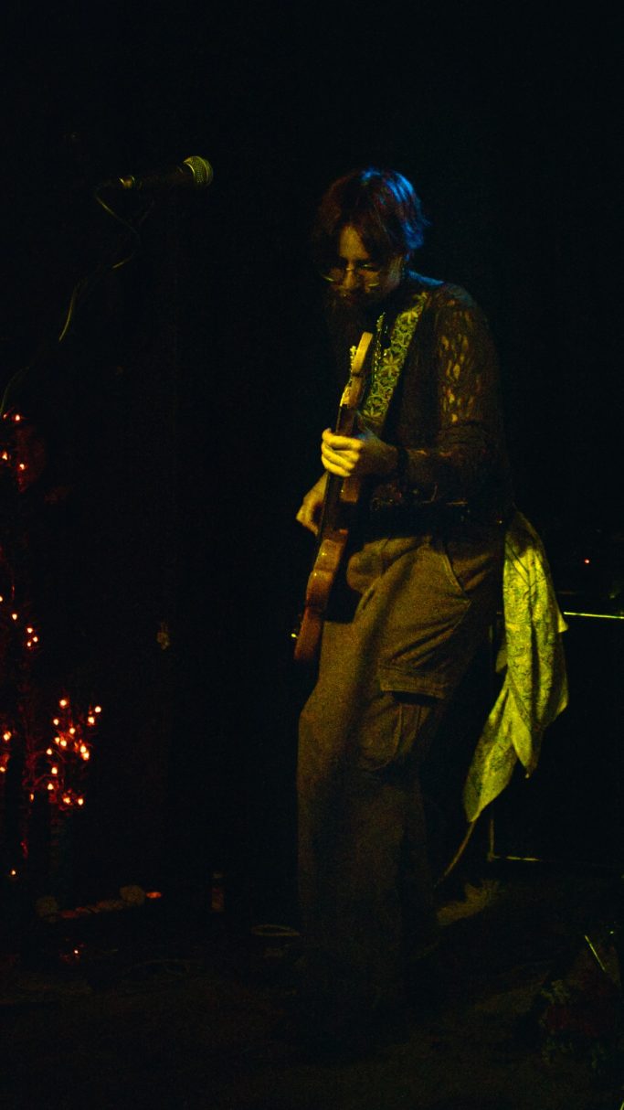 Ein Musiker spielt Gitarre auf einer dunklen Bühne mit farbigem Licht.