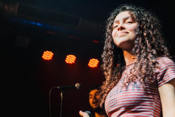 Lächelnde Frau mit lockigem Haar singt auf einer Bühne mit Bühnenbeleuchtung.
